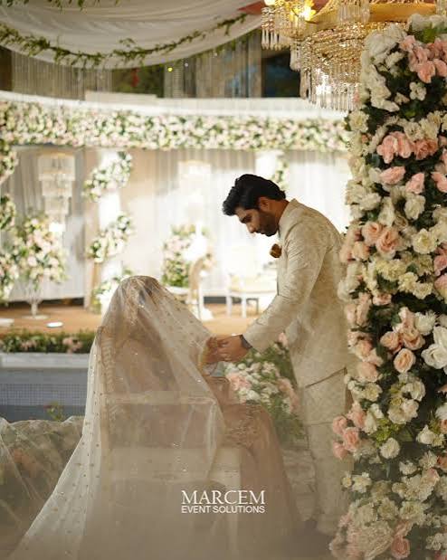 Groom in a cream sherwani greeting his veiled bride on a stage decorated with fresh roses and chandeliers